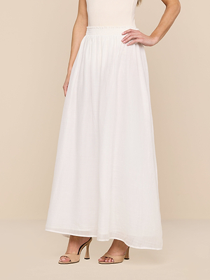 A woman stands with one hand on her hip, wearing a sleeveless beige top, beige heeled sandals, and the Evangelina Maxi Skirt—a long, breathable white skirt—against a light beige background.