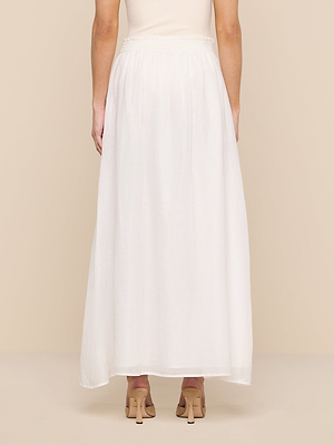 A person faces away from the camera, wearing the Evangelina Maxi Skirt—a long, white, breathable maxi skirt—and beige heeled sandals against a plain beige background.