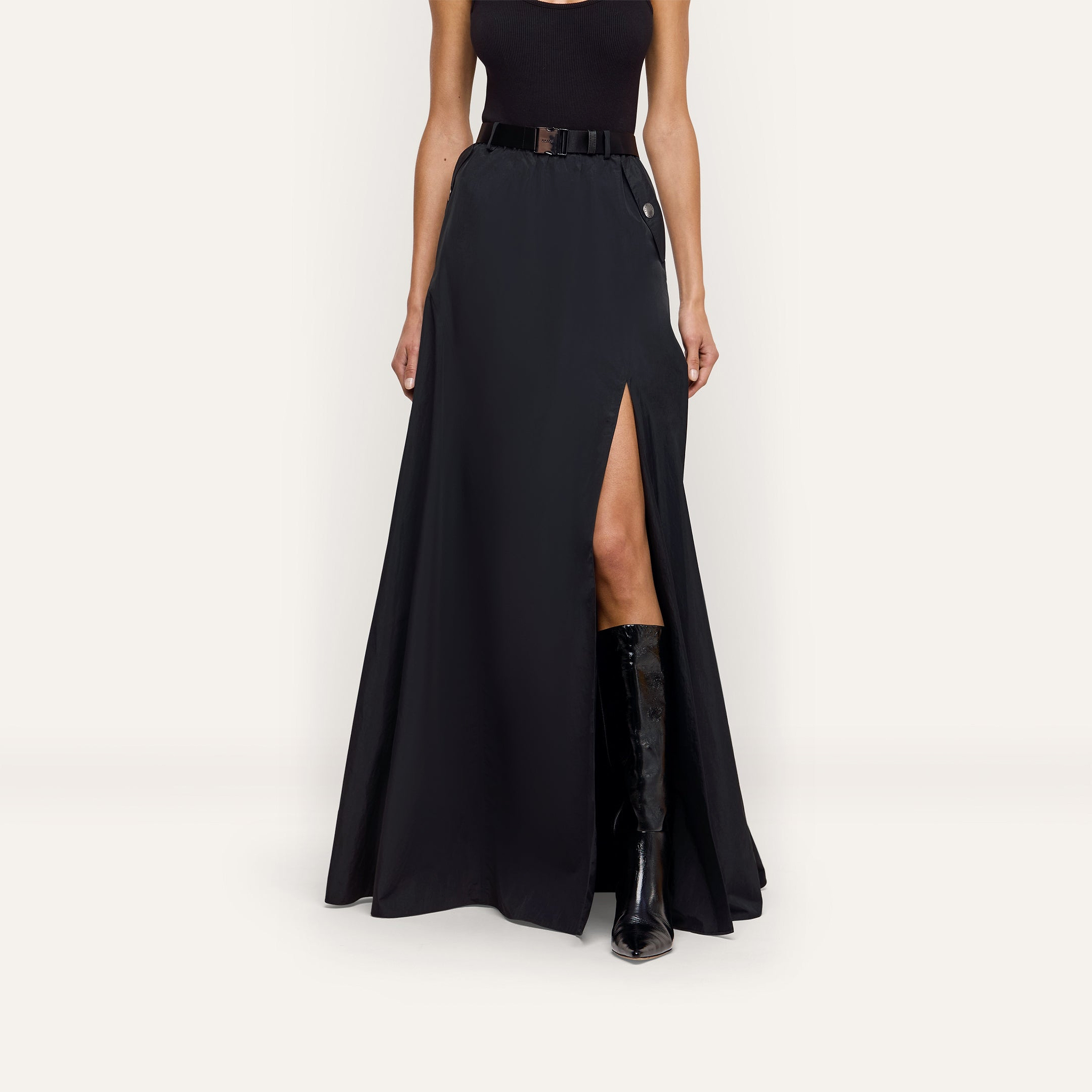 A black Sylas Nylon Skirt in a water-repellant fabric featuring a high front slit, worn with a black belt and knee-high boots.