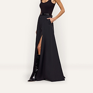 A person wears a black Sylas Nylon Skirt, a water-repellant maxi featuring a high front slit, a belt, and a visible pocket, paired with a black tank top and knee-high boots.