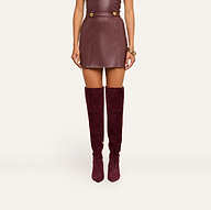 A woman wearing the Nolina Mini Skirt in mahogany brown vegan leather with gold buttons and matching knee-high suede boots, standing against a plain light background.