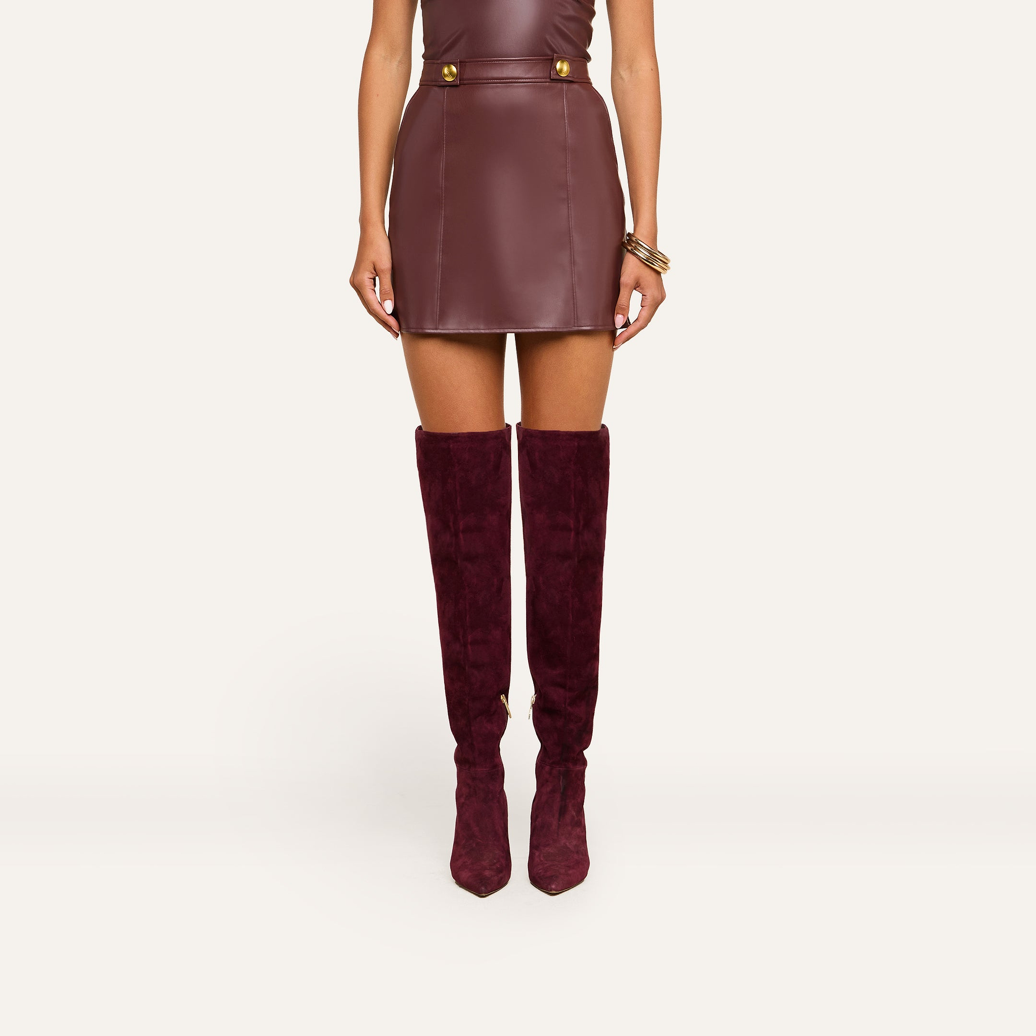 A woman wearing the Nolina Mini Skirt in mahogany brown vegan leather with gold buttons and matching knee-high suede boots, standing against a plain light background.