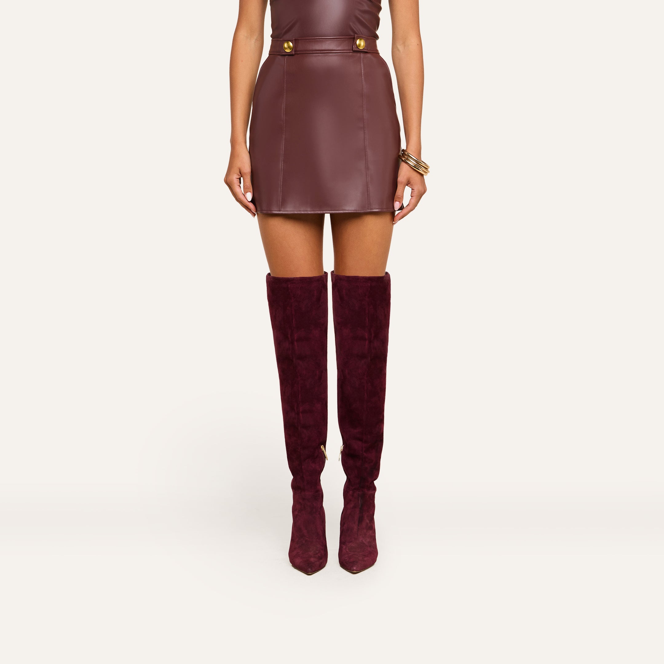 A woman wearing the Nolina Mini Skirt in mahogany brown vegan leather with gold buttons and matching knee-high suede boots, standing against a plain light background.