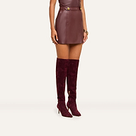 A person wearing the Nolina Mini Skirt in mahogany brown vegan leather, with a matching sleeveless top, gold bracelets, and knee-high burgundy suede heeled boots stands against a plain beige background.
