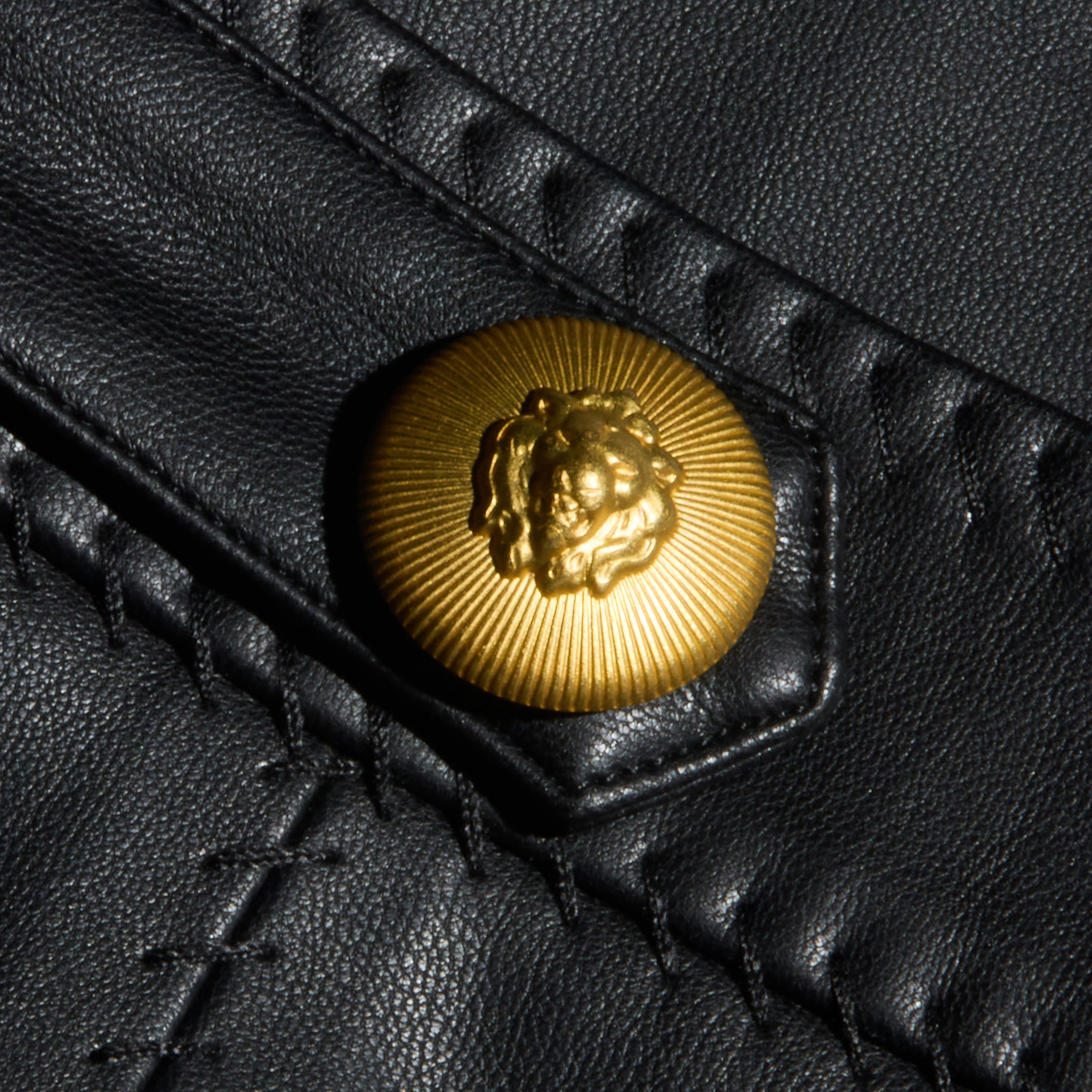 Close-up of a gold lion’s head embossed button on black Arlena Vegan Leather Mini Skirt, featuring visible whip-stitch seams and detailed stitching.
