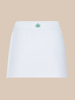 The Iga Tennis Skort in white features an A-line silhouette with a wide waistband and a small green fleur-de-lis emblem centered at the front, shown against a beige background.