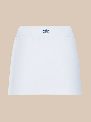 Introducing the Iga Tennis Skort: a white, high-waisted skort adorned with a small dark blue emblem at the center. Made from performance fabric with moisture-wicking properties, it delivers lasting comfort both on and off the court. Styled against a solid beige background.