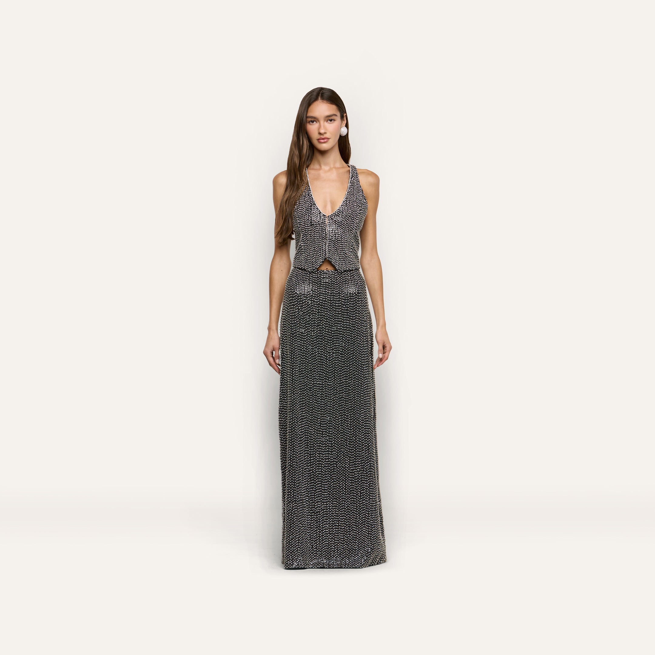 A woman wears a hand-beaded L’AGENCE sleeveless, deep-V neck top and matching Zeta Beaded Maxi Skirt, both covered in silver beading.