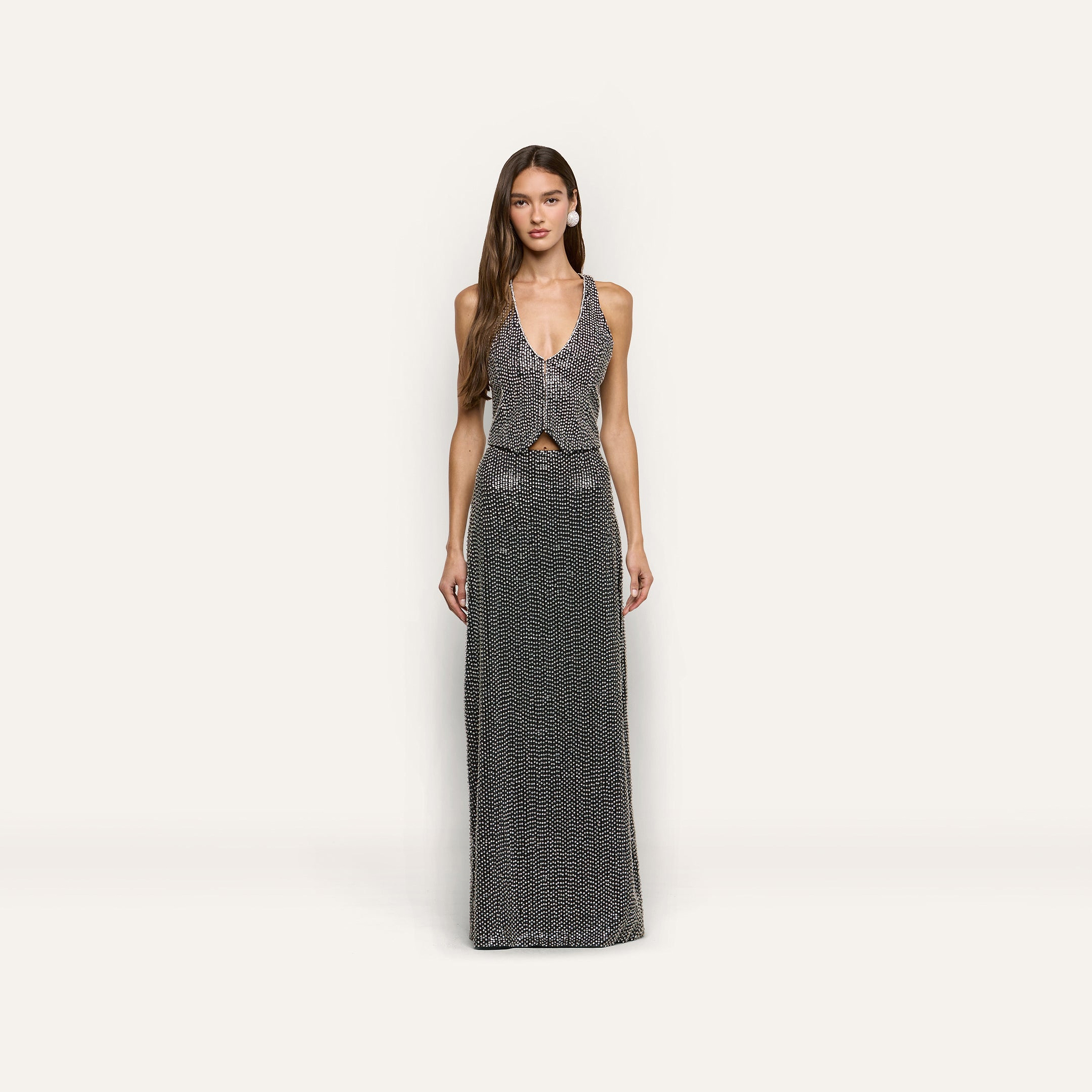 A woman wears a hand-beaded L’AGENCE sleeveless, deep-V neck top and matching Zeta Beaded Maxi Skirt, both covered in silver beading.
