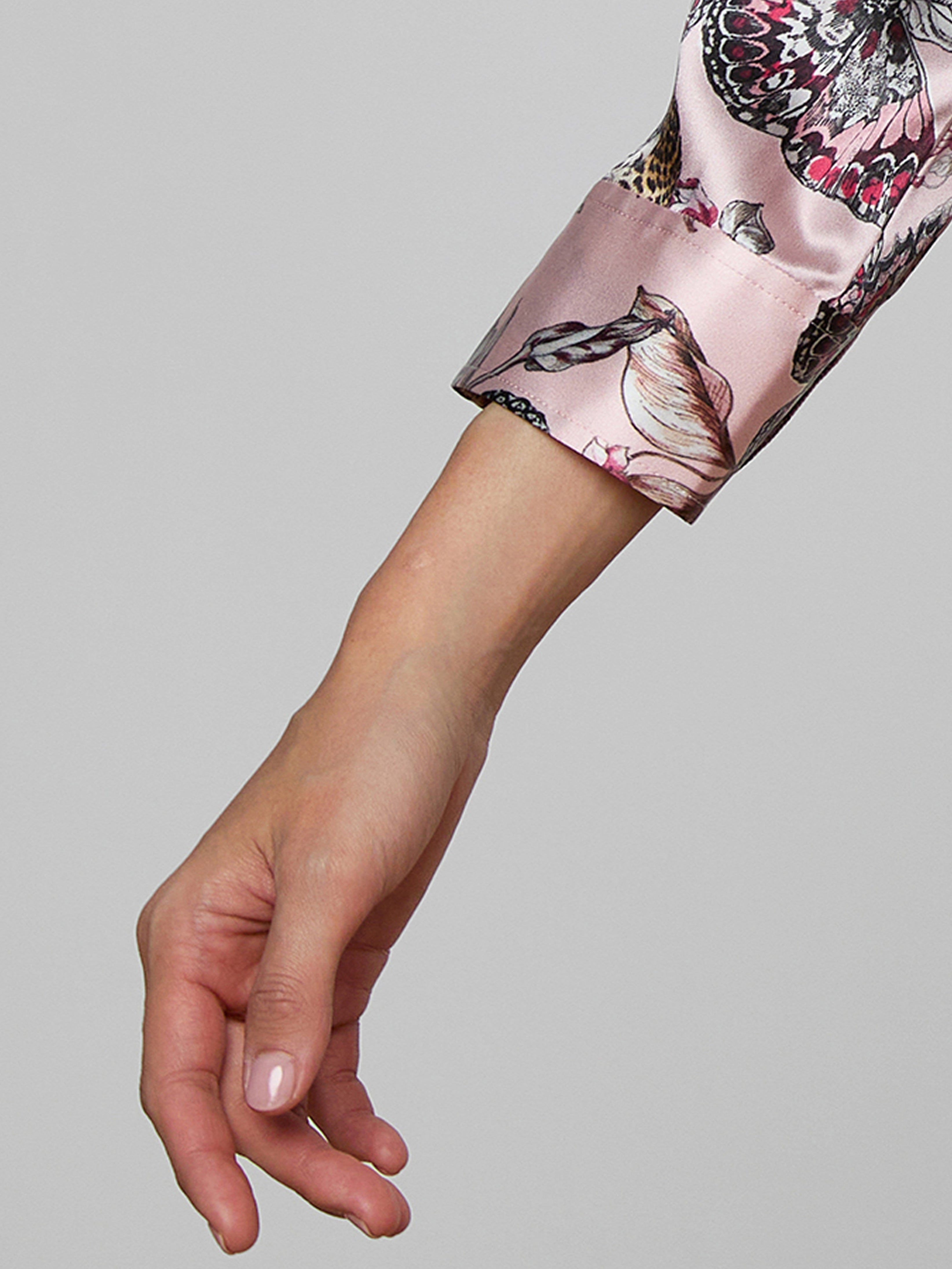 A close-up of a persons left hand and wrist wearing a long-sleeved, light pink satin shirt with a butterfly and floral pattern on the sleeve, set against a plain gray background.