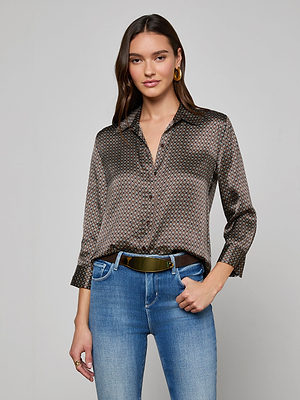 A woman with long brown hair, wearing the Dani Silk Blouse in an art deco print and blue jeans, stands against a light gray background with her left hand in her pocket and a slight smile on her face.