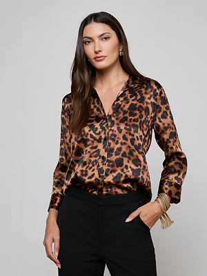 A woman with long brown hair wears the L'AGENCE Dani Silk Blouse in a leopard print with black pants, gold hoop earrings, and several gold bracelets, standing against a plain light gray background.