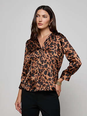 A woman with long brown hair wears the Dani Silk Blouse by L'AGENCE—a leopard print, long-sleeve shirt—paired with black pants and stands against a plain light gray background.