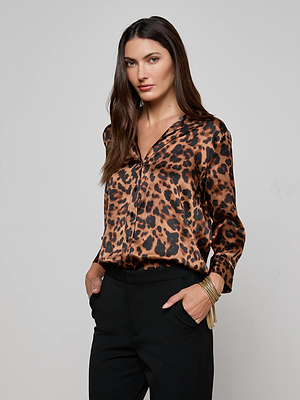 A woman with long brown hair wears the L'AGENCE Dani Silk Blouse, a leopard print button-down, paired with black pants and gold bangles, standing hands in pockets against a plain light gray background.