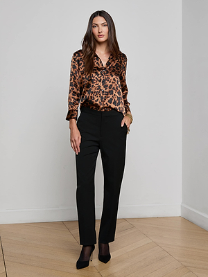 A woman stands indoors on a wooden floor, confidently looking at the camera, wearing the Dani Silk Blouse by L'AGENCE in a brown leopard print, paired with black tailored pants and black heels. She has long brown hair and one hand in her pocket.