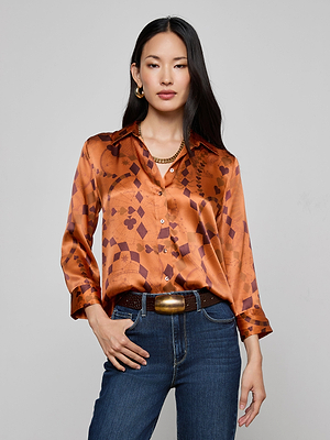 A woman with long, straight black hair wears the shiny, patterned copper-orange Dani Silk Blouse with dark blue jeans, a chunky brown belt, gold hoop earrings, and a gold chain necklace against a plain light gray background.