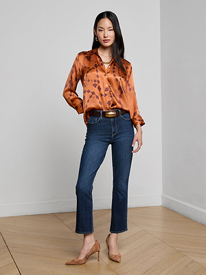 A woman with long dark hair stands indoors on a wooden floor, wearing the Dani Silk Blouse in a shiny orange pattern, paired with dark blue jeans and tan high heels. She poses against a plain background with one hand in her pocket.