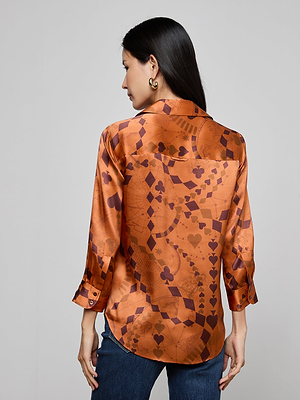 A woman with long dark hair faces away, wearing the Dani Silk Blouse—a three-quarter sleeve orange satin button-down featuring a geometric and playing card suit pattern—paired with blue jeans.