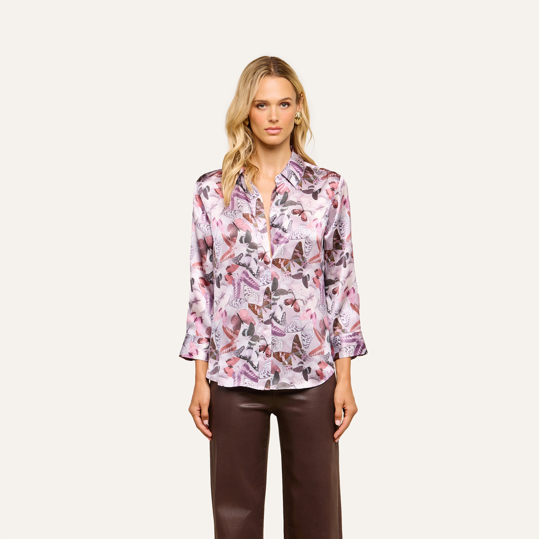 A woman wearing the Dani Silk Blouse—a silky, long-sleeve button-down featuring a mauve butterfly print—paired with brown pants, stands against a plain, light background and looks forward with a neutral expression.