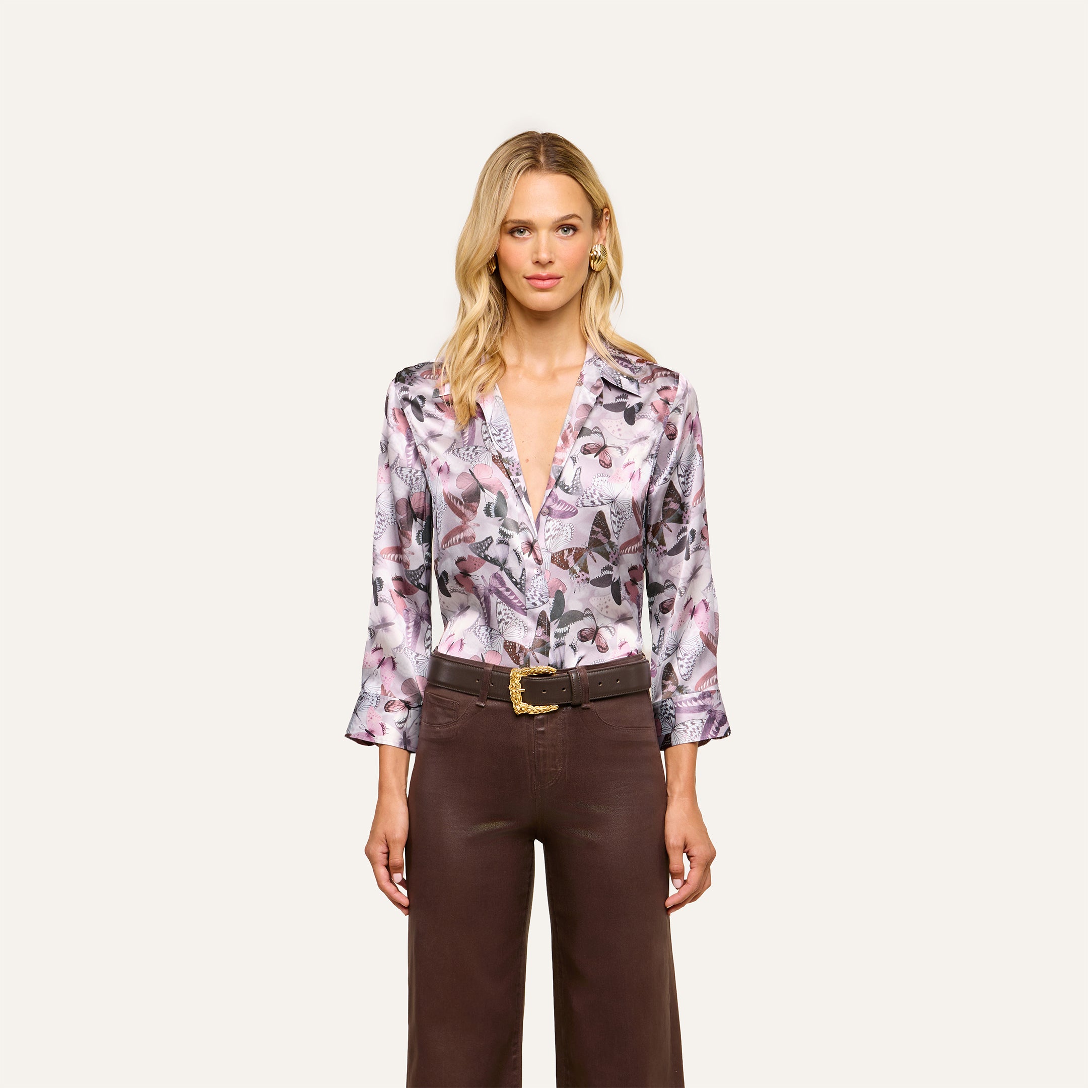 Wearing the Dani Silk Blouse in a mauve butterfly print with pink and purple tones, she pairs it with brown wide-leg belted pants and stands against a simple light background.