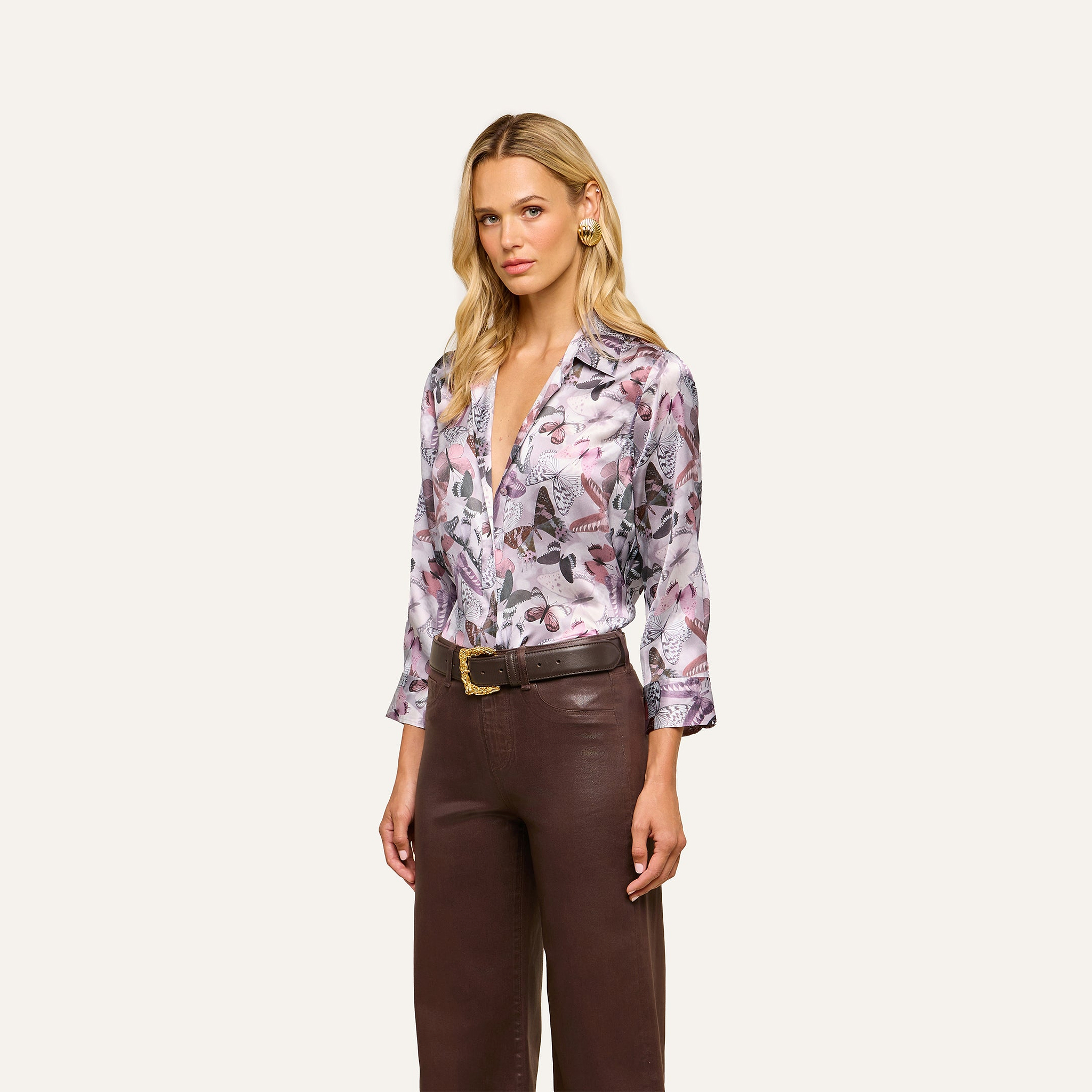 A woman with long blonde hair is wearing the Dani Silk Blouse—a button-down top featuring a mauve butterfly print—paired with high-waisted brown leather pants, standing against a plain light background.