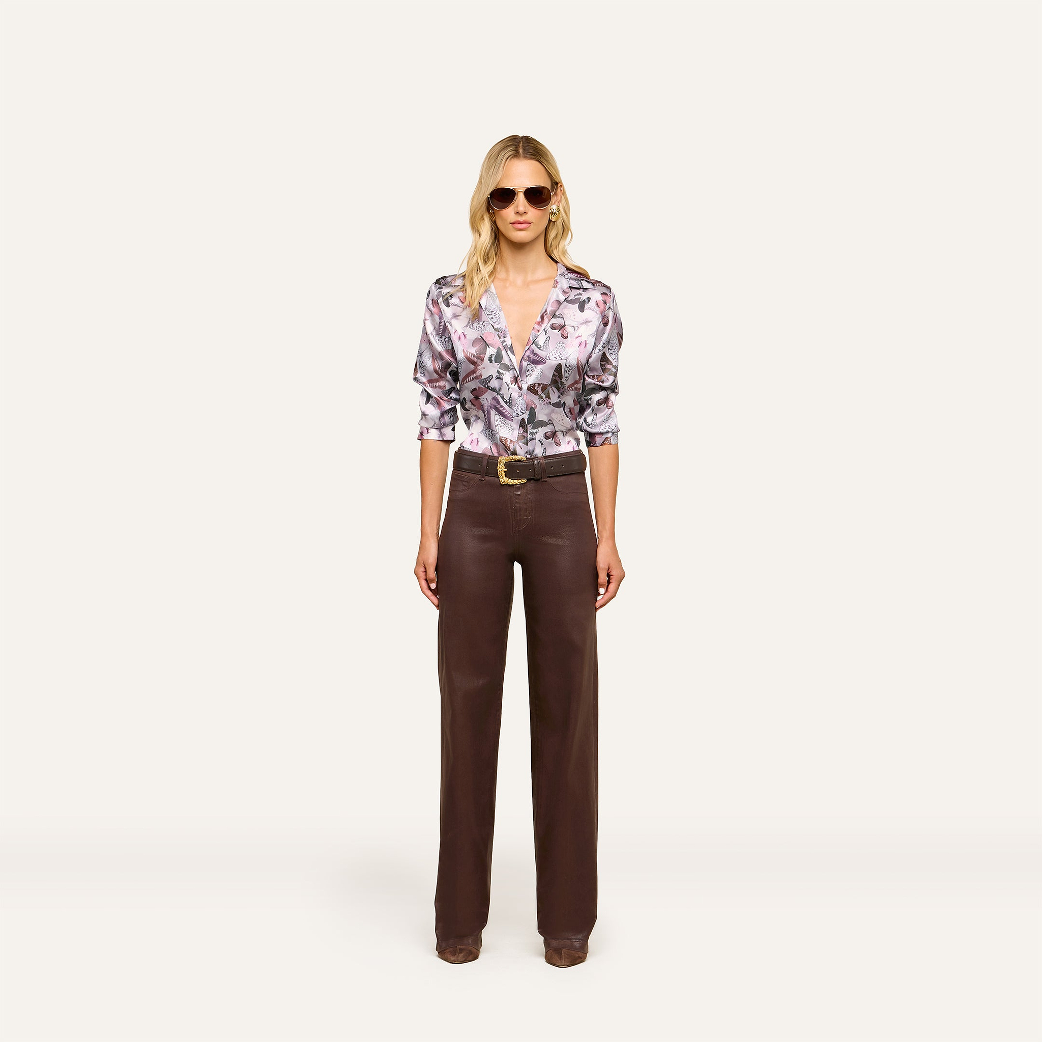 A woman wearing sunglasses, a mauve butterfly print Dani Silk Blouse, wide-leg brown trousers, and brown shoes stands facing forward against a plain white background.