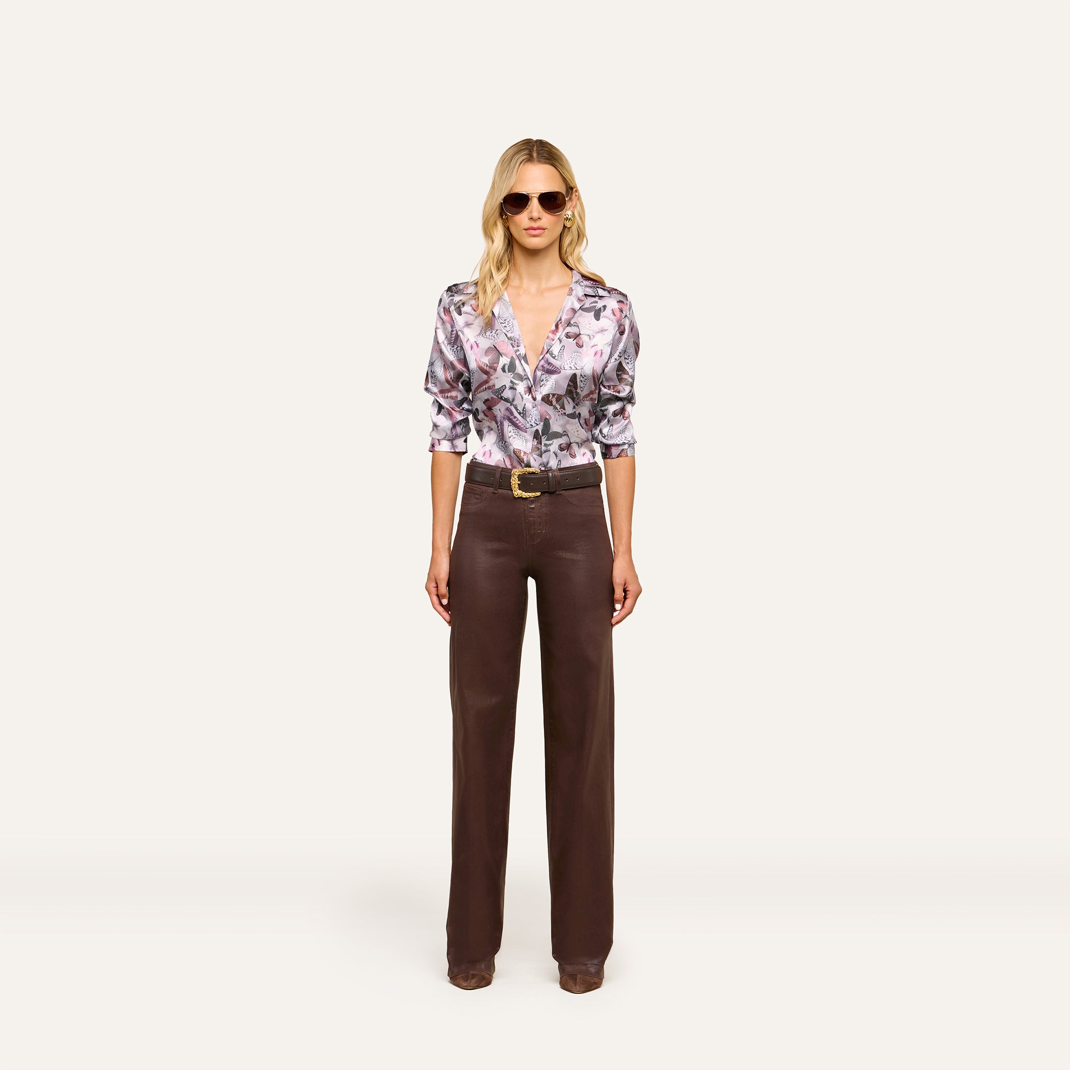 A woman wearing sunglasses, a mauve butterfly print Dani Silk Blouse, wide-leg brown trousers, and brown shoes stands facing forward against a plain white background.