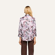 A woman with long blonde hair is shown from the back wearing a light-colored Dani Silk Blouse with a mauve butterfly print and long sleeves, paired with brown pants, standing against a plain beige background.
