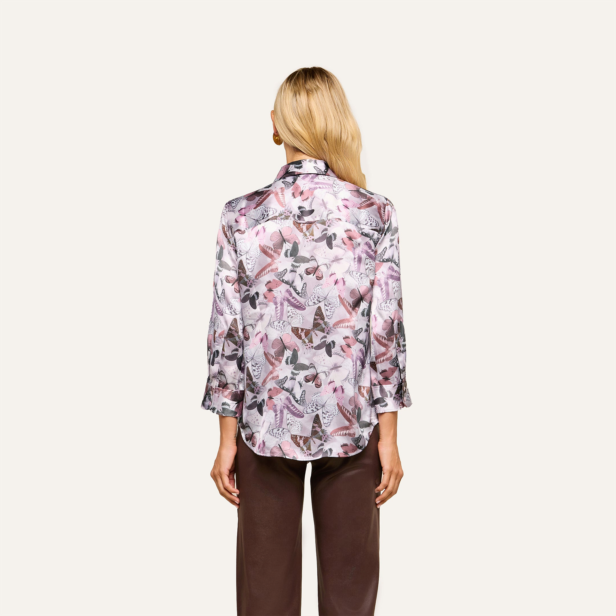 A woman with long blonde hair is shown from the back wearing a light-colored Dani Silk Blouse with a mauve butterfly print and long sleeves, paired with brown pants, standing against a plain beige background.
