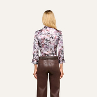 A woman with blonde hair stands facing away, wearing the Dani Silk Blouse—a long-sleeve silky button-down featuring a mauve butterfly print—paired with high-waisted brown leather pants against a simple light background.