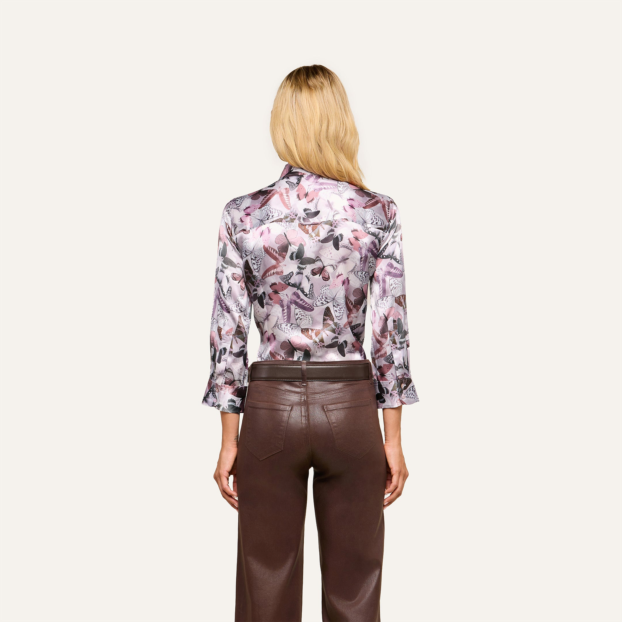 A woman with blonde hair stands facing away, wearing the Dani Silk Blouse—a long-sleeve silky button-down featuring a mauve butterfly print—paired with high-waisted brown leather pants against a simple light background.