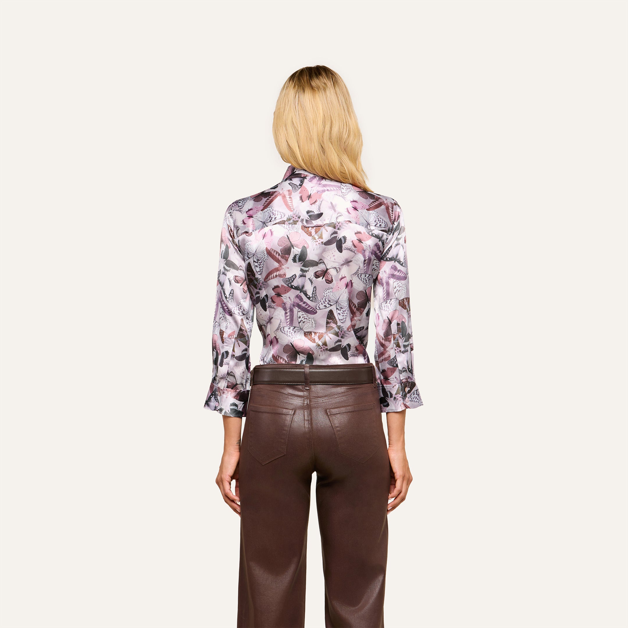 A woman with blonde hair stands facing away, wearing the Dani Silk Blouse—a long-sleeve silky button-down featuring a mauve butterfly print—paired with high-waisted brown leather pants against a simple light background.