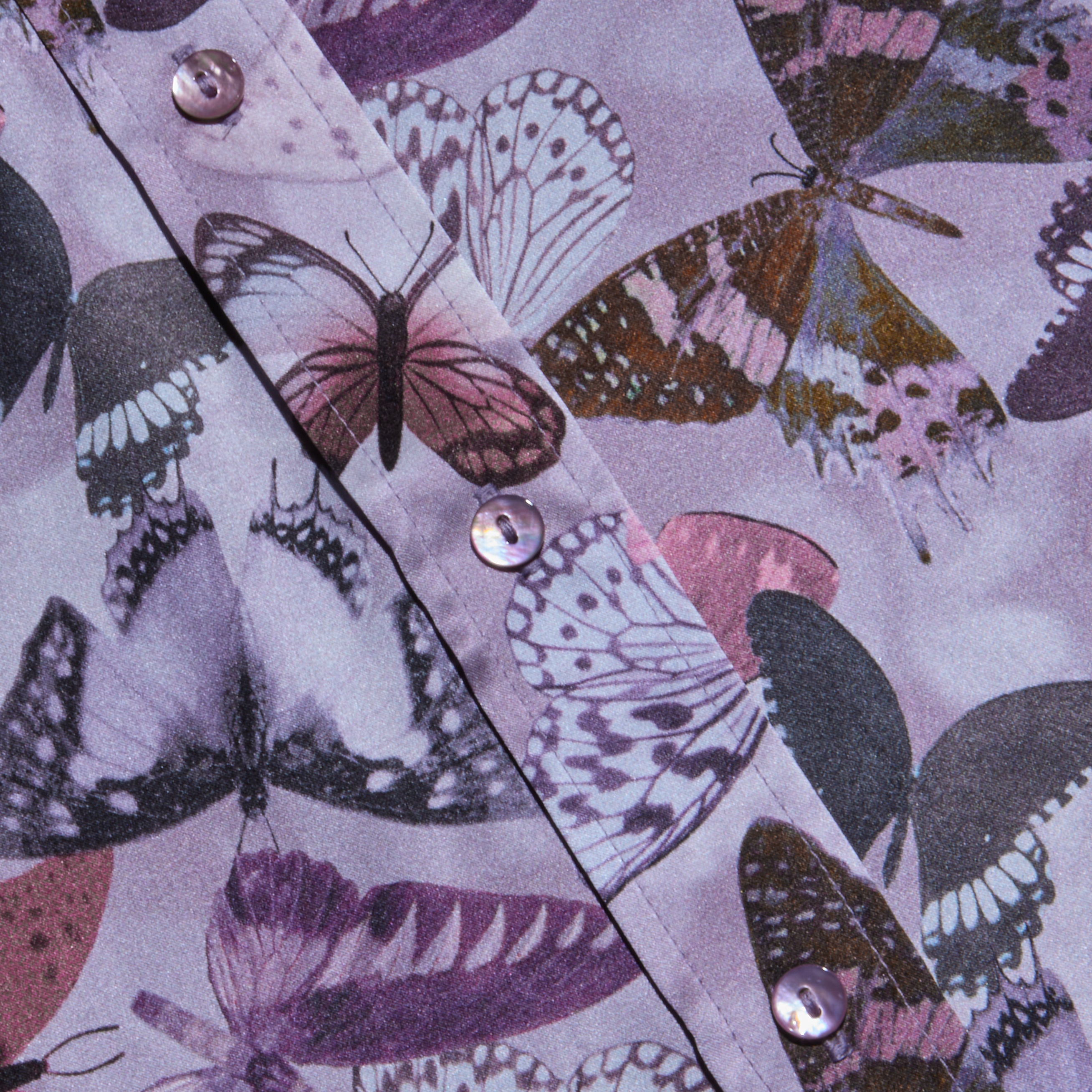 Close-up of the Dani Silk Blouse, featuring a mauve butterfly print with matching buttons. The elegant button-down showcases various butterfly species in purple, black, and white on luxurious silk.