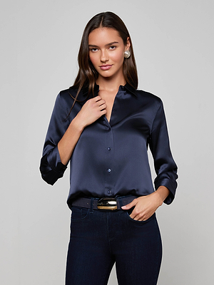 A woman with long brown hair wears the midnight blue Dani Silk Blouse by L'AGENCE, paired with dark jeans and pearl earrings. She stands against a light background, gently touching her collar and smiling slightly.
