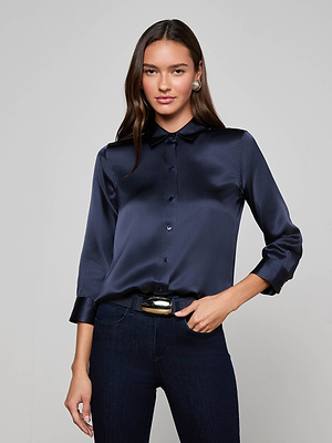 A woman with long brown hair wears the L'AGENCE Dani Silk Blouse in midnight blue and dark jeans, standing against a light gray background with one hand on her hip and a neutral expression.