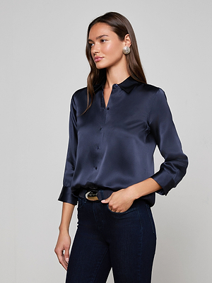 A woman with long brown hair models the L'AGENCE Dani Silk Blouse in midnight blue, featuring three-quarter sleeves, paired with dark jeans. She stands against a light gray background, gazing to the side with one hand in her pocket.
