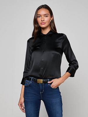 A woman models the L'AGENCE Dani Silk Blouse in black with long sleeves, paired with blue jeans, as she stands against a light gray background and smiles slightly at the camera.