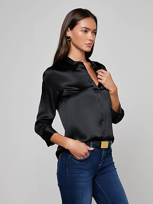 A woman with long brown hair wears the L'AGENCE Dani Silk Blouse in black, dark blue jeans, a black belt with a gold buckle, and gold earrings, standing against a plain light gray background.