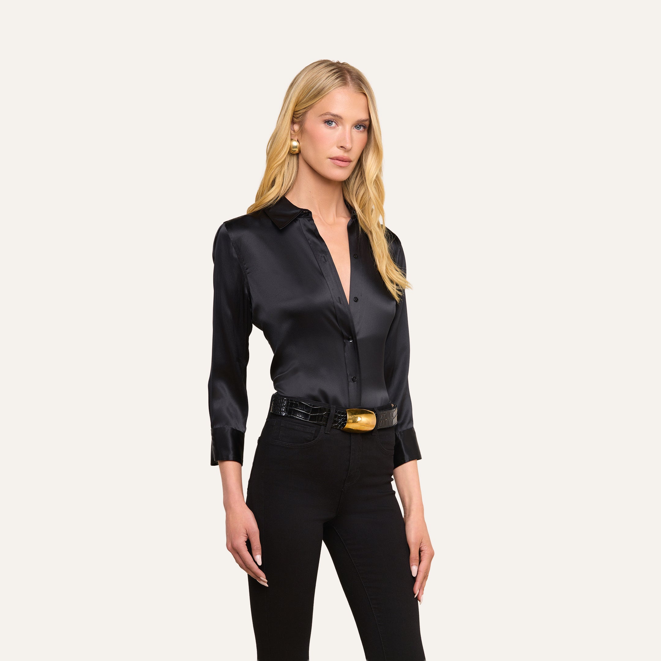 Una donna dai lunghi capelli biondi indossa la camicetta Dani Silk di colore nero, abbinata a pantaloni neri, una cintura nera con fibbia dorata e orecchini dorati, in piedi su uno sfondo beige.