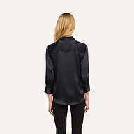Una donna dai lunghi capelli biondi, vista di spalle, indossa la camicetta Dani Silk nera a maniche lunghe e pantaloni neri, in piedi su uno sfondo beige a tinta unita.