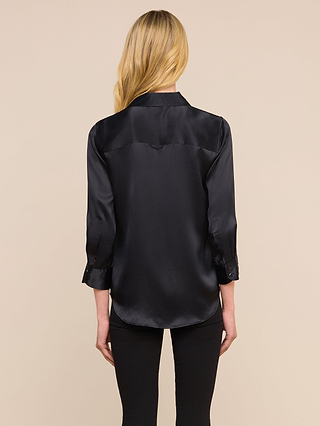 Une femme aux longs cheveux blonds, vue de dos, porte le chemisier « Dani Silk » noir à manches longues et un pantalon noir, debout devant un fond beige uni.