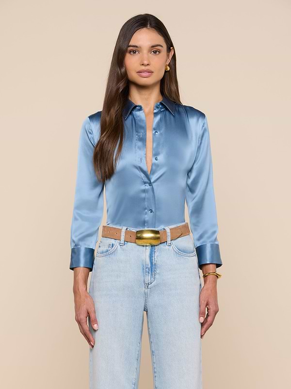 A woman with long brown hair wears a shiny blue-mist Dani Silk Blouse tucked into light blue jeans with a tan belt, standing against a plain light background.