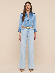 A woman stands facing forward against a plain white background, wearing the shiny blue mist Dani Silk Blouse tucked into light-wash high-waisted wide-leg jeans, styled with a beige belt and tan heels.
