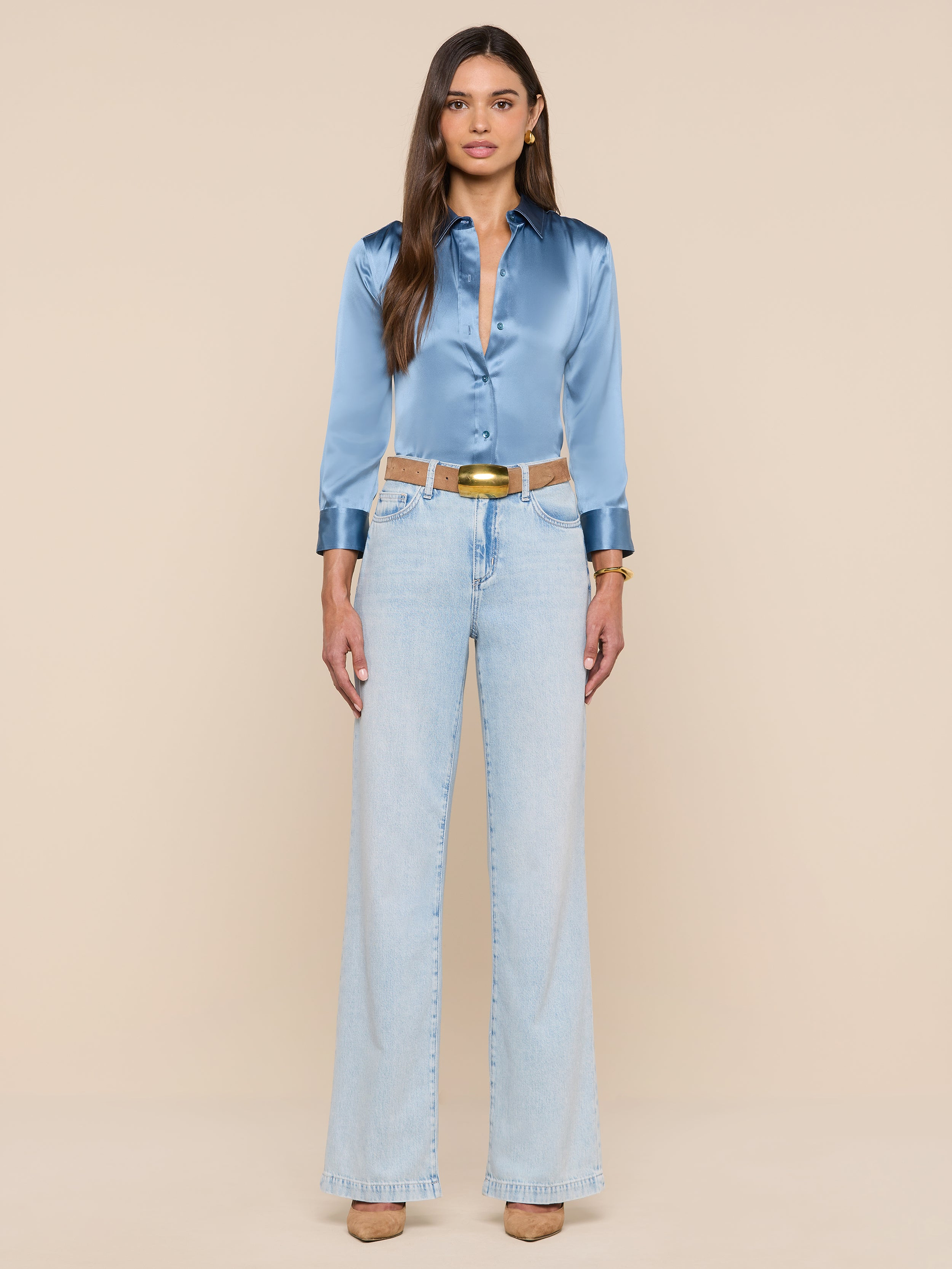 A woman stands facing forward against a plain white background, wearing the shiny blue mist Dani Silk Blouse tucked into light-wash high-waisted wide-leg jeans, styled with a beige belt and tan heels.