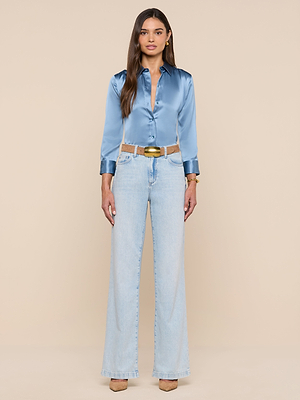 A woman with long brown hair and a neutral expression stands against a beige background wearing a shiny blue mist Dani Silk Blouse, light blue high-waisted wide-leg jeans, a tan belt, and beige heels.
