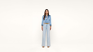 A woman stands against a plain white background, wearing a light blue satin blouse, high-waisted wide-leg jeans, and beige heels.