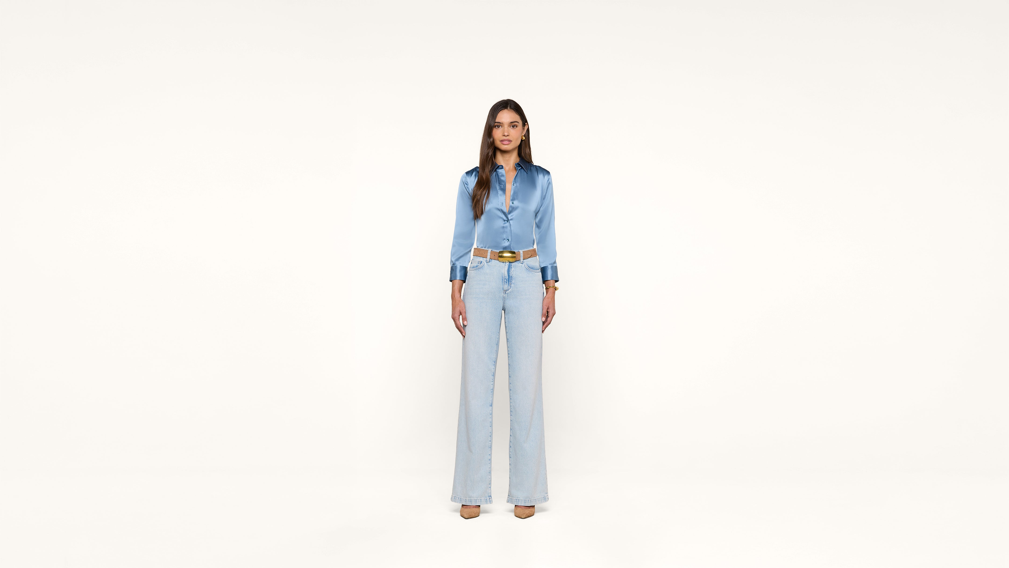 A woman stands against a plain white background, wearing a light blue satin blouse, high-waisted wide-leg jeans, and beige heels.