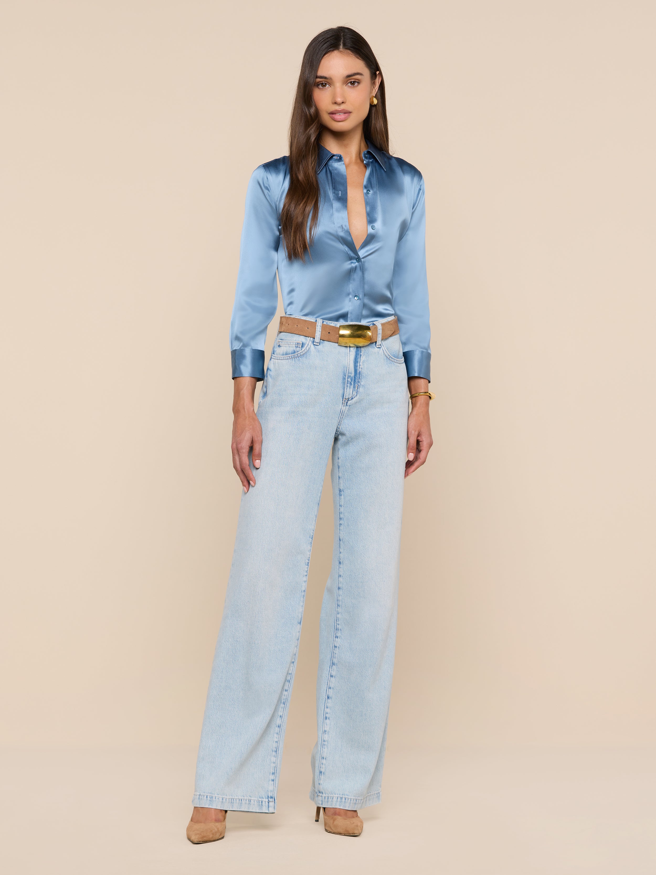 A woman in a blue mist Dani Silk Blouse and light blue high-waisted jeans stands against a plain white background, wearing tan heels, a white belt, and a gold watch.