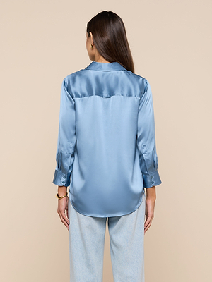 A woman with long brown hair is seen from behind, wearing a shiny blue mist Dani Silk Blouse with long sleeves and light blue jeans, standing against a plain beige background.
