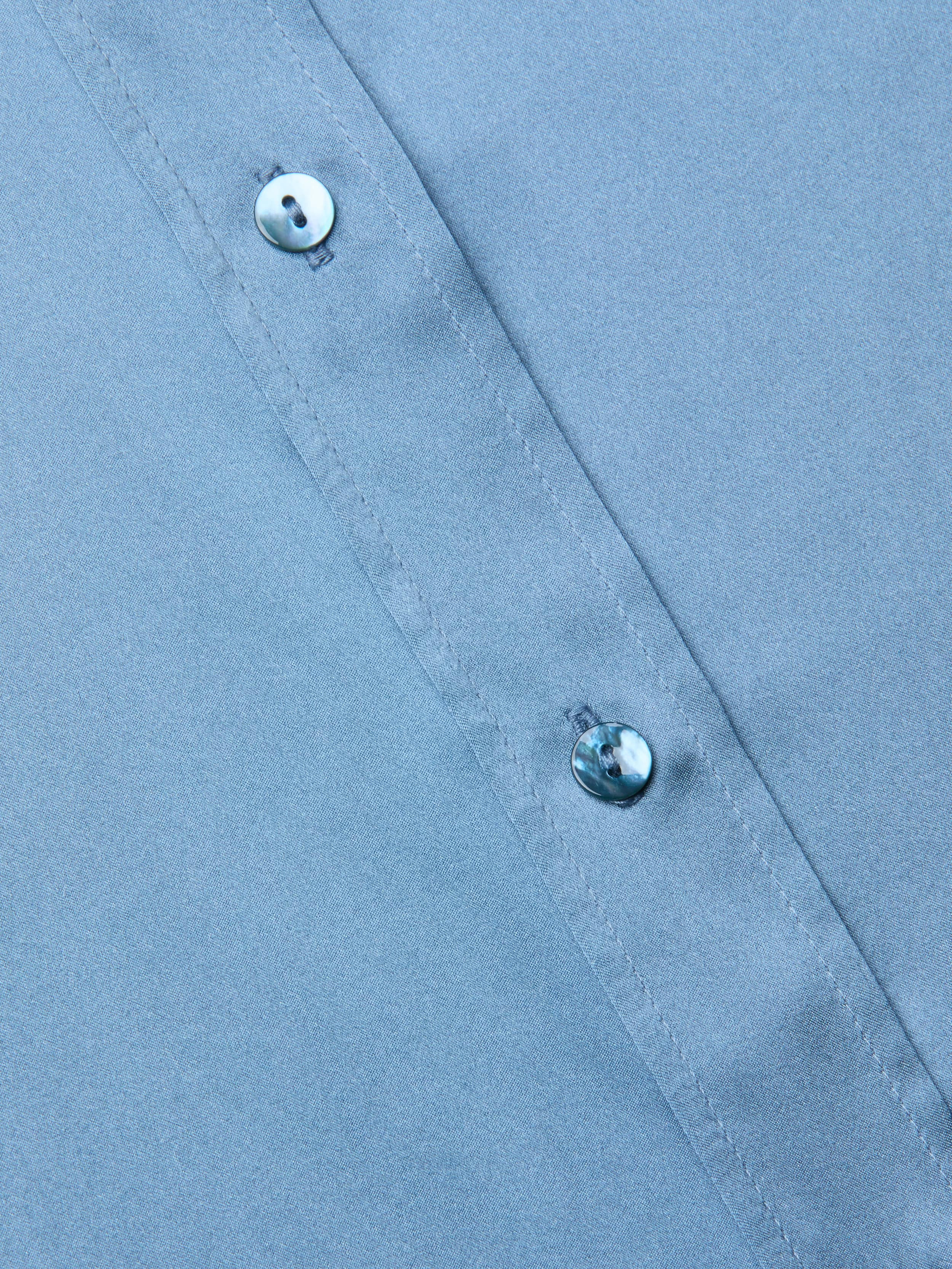 Close-up of a blue mist Dani Silk Blouse fabric with two small, shiny buttons along a stitched seam, shown at a slight diagonal angle.